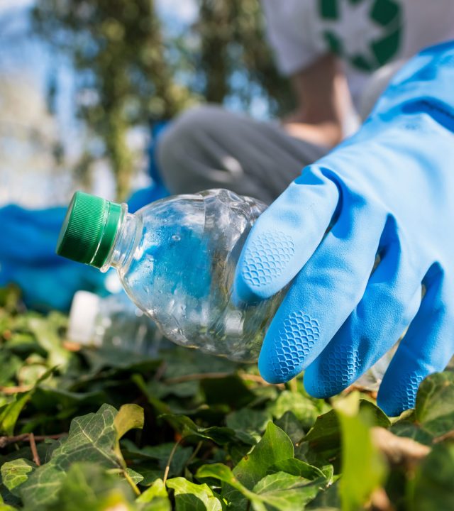 Man collecting plastic garbage in container in a polluted park. Rubber gloves, recycling signs on T-shirt